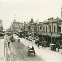 Image: a dirt street, lined with two and three storey stone buildings and busy with horse drawn vehicles, pedestrians and street vendors. 