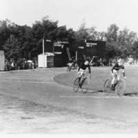 Image: cycle racing at Adelaide Oval. Two men on bicycles vie for the lead on a tarmac track.