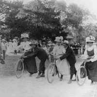 Image: three women on bicycles about to begin a race. Men hold the bicycles steady.