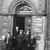Image: A group of men in early twentieth century attire stand in front of a large, nineteenth century two-storey bluestone building. Signs in the first floor windows read ‘Seamen’s Mission & Sailors’ Rest’ 