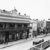 Image: Shops and pedestrians seen from an elevated position on the opposite side of the road. The row of two storey shops in the centre has parapet and a cantilever verandah. The building on the left is covered in scaffolding.