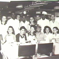 Image: Nineteen men and women in a ship's kitchen. Seven women and one man are seated at a table, the rest stand behind them. They are all wearing white or aprons