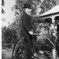 Image: man in a top hat and suit seated on a penny-farthing bicycle