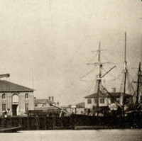 Image: A mid-nineteenth century landscape photograph of a port waterfront. Several buildings are visible next to the water, and a small number of sailing ships are moored alongside a large wharf