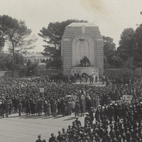 Image: A large crowd of people in 1930s dress or military uniforms gather around a huge stone monument featuring three bronze statues of a girl, a student and a farmer in front of a large marble angel.