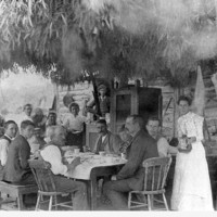 Image: Men and Women around a table underneath a brush and eucalyptus shelter