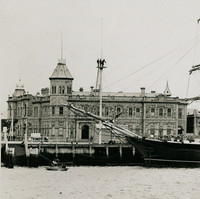 Image: A large, three-masted sailing ship is moored alongside a wharf in a port town. Several buildings, including a large nineteenth-century structure with an octagonal tower in one corner, are visible in the immediate background