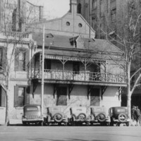 Image: 1930s era cars are parked on the street outside a two-storey stone building with shuttered windows and a balcony running halfway along the front of the building. 