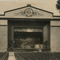 Image: a large open air theatre with rows of seats facing a stage with an outdoor scene as its backdrop