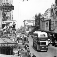Image: 1920s and 1930s era cars, men on bicycles and a double decker bus travel down a busy city street lined with a large number of shops and a theatre
