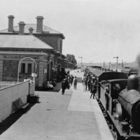 Image: A railway station. A train is on the platform to the right, with smoke coming out of the front. People are boarding the train. To the left is the station building, made of brick