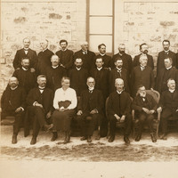 Image: Black and white group portrait. Three rows of men in suits pose against a stone building. There is one woman in the photograph. She is seated in the front row, third from left.