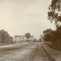 Image: a wide, muddy road stretches through parklands. In the distance the road continues through a more built-up area with numerous buildings including a corner hotel.