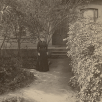 Image: Black and white photograph of a woman in front of a house