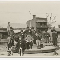 Image: A group of children sit on a merry-go-round (or joy wheel) in a park. A street and row of buildings are visible in the background.
