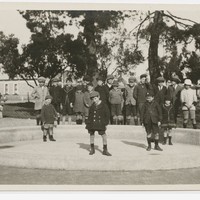Image: A group of children pose for a photograph in an empty wading pool in a park. A street and row of buildings are visible in the background.