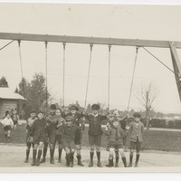 Image: A group of children pose for a photograph under a swing in a park. A building and trees are visible in the background