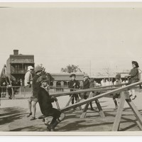 Image: A group of children play on see-saws in a park. A street and row of buildings are visible in the background