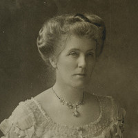 Image: Photographic portrait of a young Caucasian woman in Edwardian-era attire. She is seated and wearing a floor-length light-coloured dress with embroidered decoration