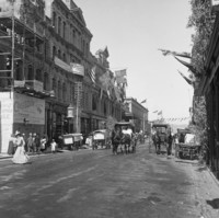 Image: A wide dirt road with a number of pedestrians and horse drawn vehicles. The buildings are decorated with flags and greenery. One of the buildings on the left is still under construction and is covered in scaffolding. 