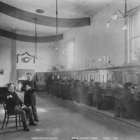 Image: Interior view of a telephone exchange. A row of women switchboard operators are supervised by a well dressed woman sitting at a raised desk on the left.