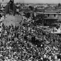 Image: A large group of people dressed in 1940s attire gather on the approach to a bridge. Several buildings are visible just beyond the bridge approach