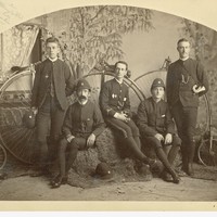 Image: a studio group of five members of the South Australia Bicycle Club with penny farthing bicycles; they are wearing the club uniform with helmet hats, one carries a small bugle on a shoulder belt. 