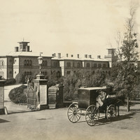 Image: a horse drawn carriage is pulled up alongside large gates. Beyond the gates a long drive curves around to meet a large two storey stone building with arched windows and a large number of chimneys.