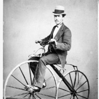 Image: man in a top hat and suit riding a velocipede with wooden wheels