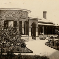 Image: a small number of women and girls in 1860s era clothing stand outside a single storey bluestone house with curved wall, columned portico and verandah.