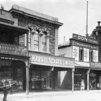 Image: a row of two storey shops with verandahs line a dirt road. To the right a horse drawn cart stands outside a hotel with a balcony.