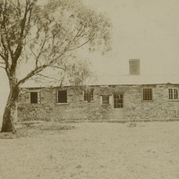 Image: Front view of a long stone building with barred windows and a tree in front. 