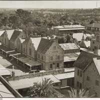 Image: an aerial view of a complex of one and two storey stone buildings with slate and tin roofs including one with verandahs under which 1930s cars are parked. 