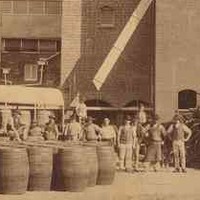 Image: a group of men in late 19th century worker's clothes stand with a collection of large barrels outside of a tall red brick brewery building.