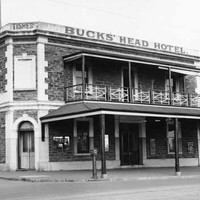 Image: a two storey corner hotel with a small balcony on one side. A verandah protrudes further out to the street beneath the balcony. A parapet sign reads: "TISHERS BUCKS' HEAD HOTEL"