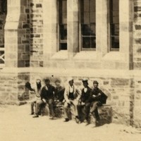 Image: A large, three-storey mid-nineteenth century bluestone building sitting atop reclaimed land. A sign reading ‘Mercantile Marine Office’ is visible at the bottom right of the building. A group of men sit on a bench near the building entrance