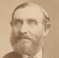 Image: A photographic head-and-shoulders portrait of a bearded middle-aged man in a suit with bowtie