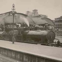Image: a steam train is stopped beside a platform on which stands a number of men and women in early 20th century clothing. Its carriages stretch away into the distance and pass under a curved roof platform cover. 