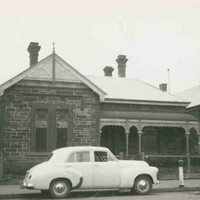 Image: a white 1950s era car is parked outside a small bluestone cottage with a verandah
