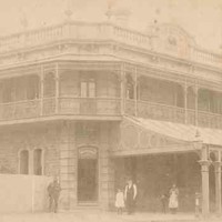Image: a two storey corner hotel with a narrow wrap around balcony and balustraded parapet with urn decorations. On one side of the ground floor a wide verandah protrudes beyond the balcony above.  The other side is fenced off. 