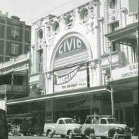 Image: 1950s era cars are parked outside a large theatre building which features a huge decorative arch inside which is a sign with the theatre's name and information about the current show. The building also features decorative plasterwork.