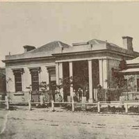 Image: a single storey building with grand columned portico under which figures can be seen standing. A garden can be seen behind a fence to the right of the building. 
