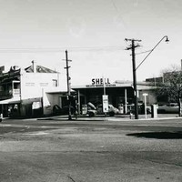 Image: A 1960s era shell service station, with a car with its bonnet up parked under the overhanging roof, sits on the corner of two city streers