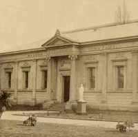 Image: Old photograph of a single storey building with a door and six window along the front and a single window visible on the adjacent side of the building.  The building also features a small portico and is reasonably ornate. 