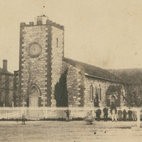 Image: A low, squat church building stands at the intersection of two dirt roads. A man stands in front of the fence bordering the churchyard