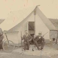Image: Four men sit on chairs in front of a tent. One man holds a bicycle while two others brandish rifles