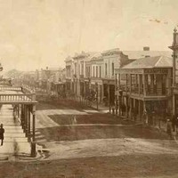 Image: a view of a city street which is lined with two and three storey commercial buildings. A large number of male and female pedestrians in 1860s attire can be seen walking down the sidewalks or crossing the road. 