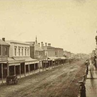 Image: a view of a city street lined with two storey commercial buildings, many of which on one side of the street have verandahs or balconies while most on the other side have flat facades. On the left, a large model of an emu stands above a verandah.