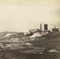 Image: Several large chimneys and buildings are arrayed among numerous tailings piles. A low mountain range is visible in the background