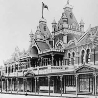 Image: a two storey ornate brick building with arched windows, a series of store windows beneath a verandah, two towers with square cupolas and flagpoles flying Australian flags above the main entrance and a tiled roof